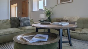 Ein Wohnzimmer mit einem grünen Sofa, einem runden Tisch mit Tablett und Büchern, einer Blumenvase und einem gerahmten Bild an der Wand.