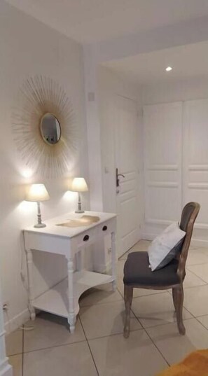 A room with a white vanity, two lamps, a mirror, a wooden chair, and a door.