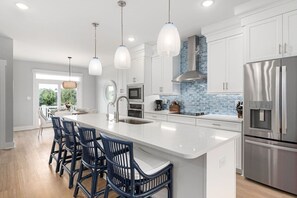Une cuisine moderne dotée d’un grand îlot central, de meubles blancs, d’appareils électroménagers en acier inoxydable et d’un dosseret de crédence recouvert de carreaux bleus.