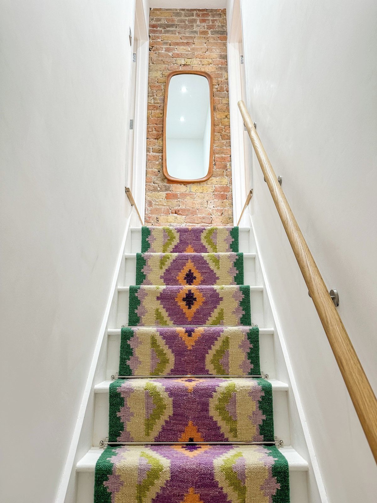 A staircase with a patterned carpet, a wooden handrail, and a brick wall with a mirror at the top.