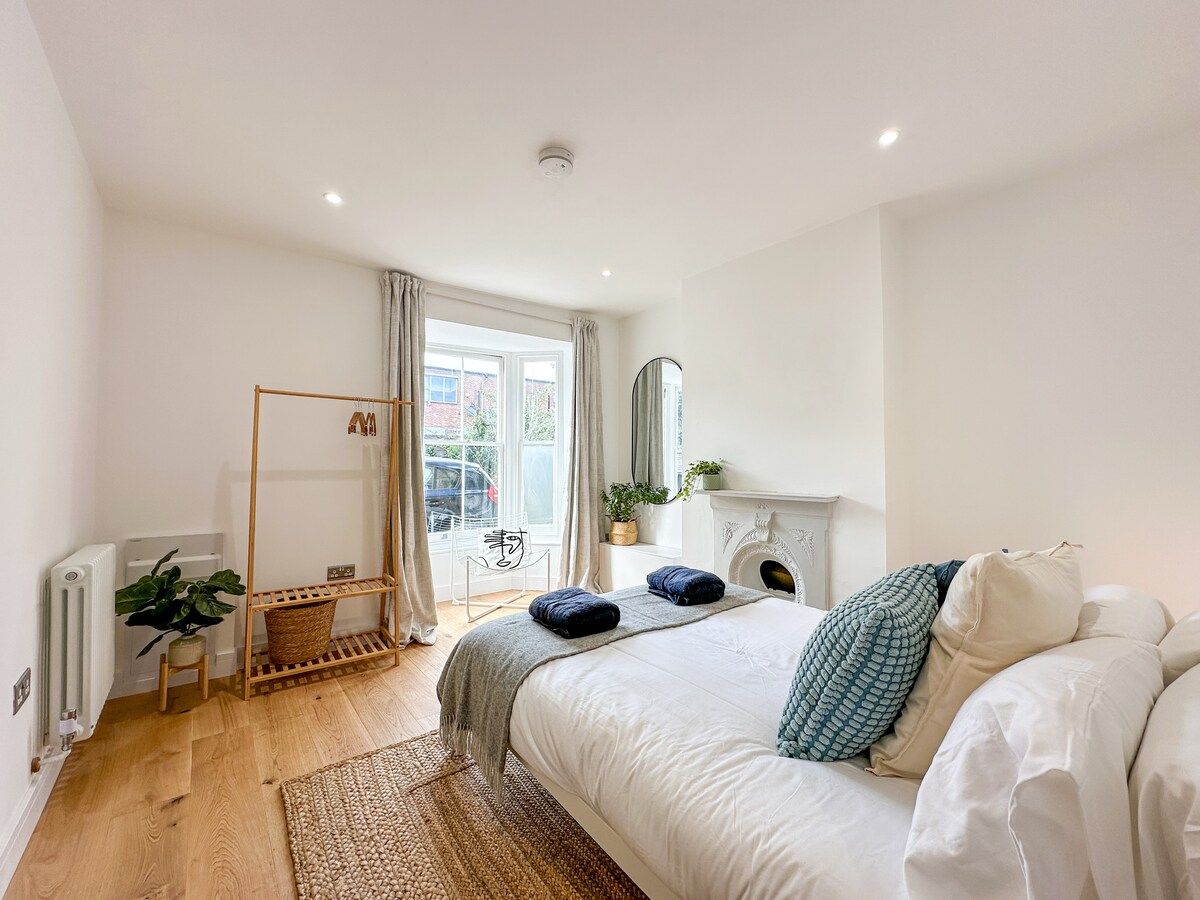 Ein Schlafzimmer mit einem Bett, einem Heizkörper, einem Fenster mit Vorhängen, einer Pflanze und einem Spiegel an der Wand.