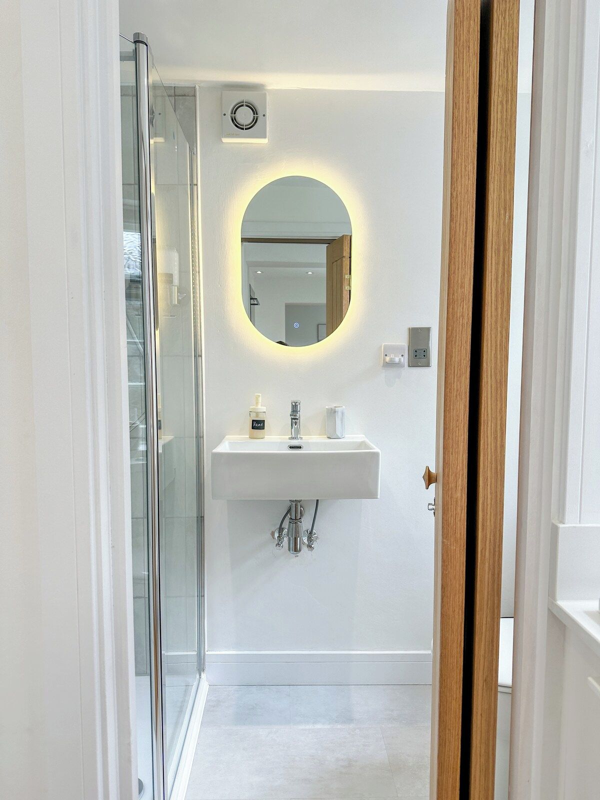 A modern bathroom with a glass shower enclosure, a white sink with a round mirror, and a wooden door.