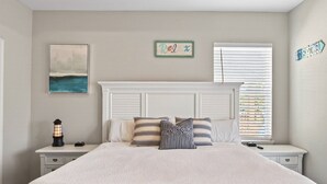 A bedroom with a bed, nightstands, a lamp, a painting, a 'Relax' sign, and a window with blinds. - Steps to the Beach, Stunning Sunrises and Sunsets! (Galveston)