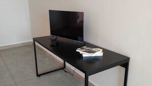 Una habitación con un televisor negro sobre un soporte, una mesa negra con un libro, y piso de baldosas.