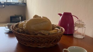 Una cesta de mimbre con pan, una tetera rosa y una taza blanca sobre una mesa de madera.
