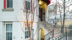 A white building with a staircase leading to a second floor, a tree in the foreground, and a fence around the property. - Chic Downtown Denver Retreat | Walk Everywhere + Stylish Comfort (Denver)
