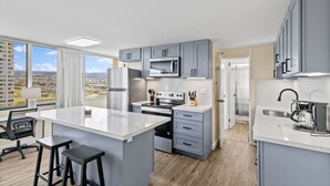 A modern kitchen with grey cabinets, a central island, and a view of a cityscape through large windows. - Skyline Access w/ Pearl Views - W/D, Parking, A/C (Pearl City)