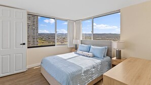 A bedroom with a large bed, a nightstand, a lamp, and a view of a cityscape through large windows. - Skyline Access w/ Pearl Views - W/D, Parking, A/C (Pearl City)