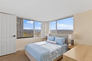 Un dormitorio con una cama grande, una mesita de noche, una lámpara y vistas a la ciudad a través de amplias ventanas.