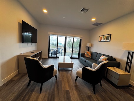 A modern living room with a flat-screen TV, a sofa, two armchairs, a coffee table, and a sliding glass door leading to an outdoor area.