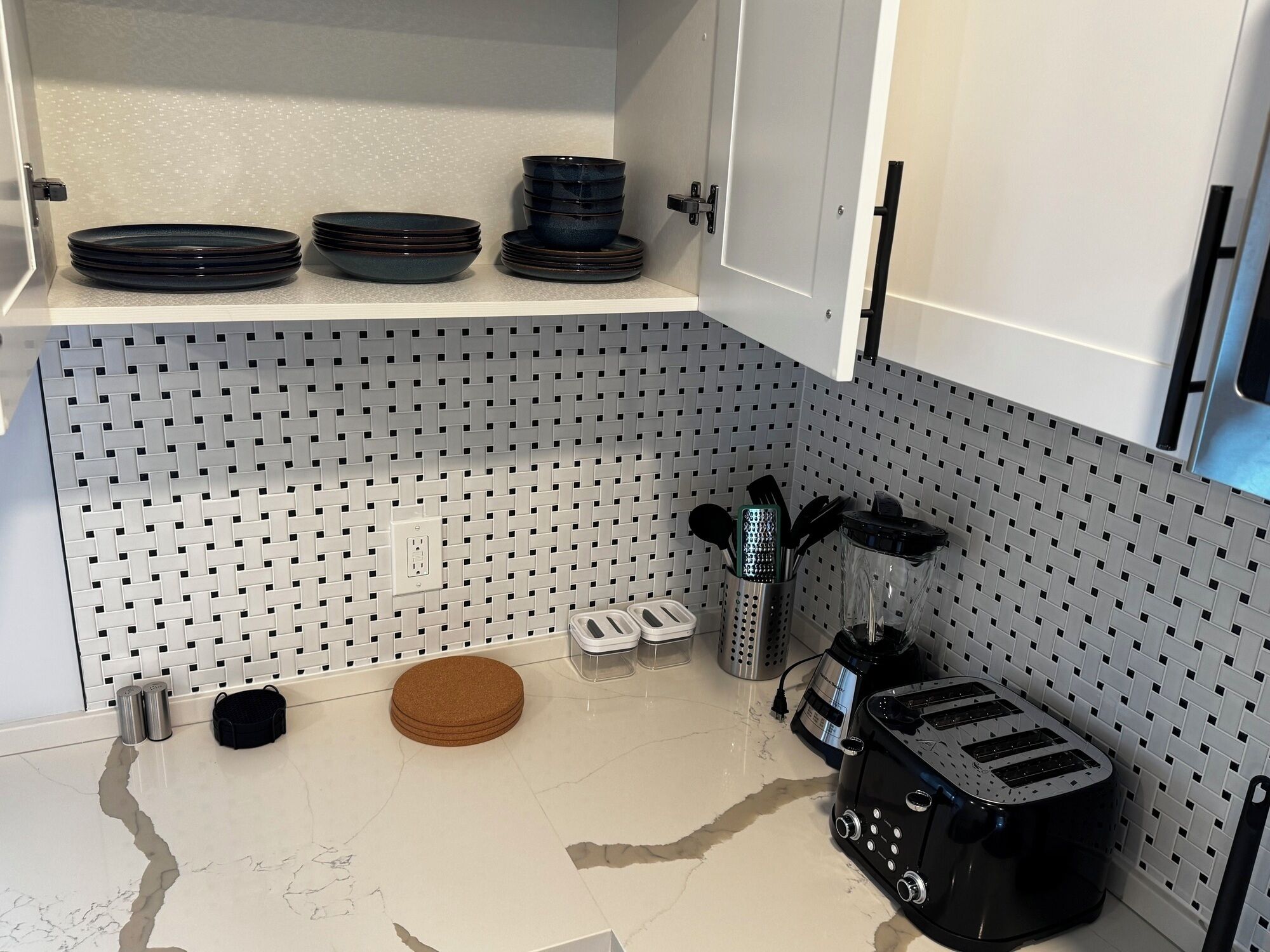 Uma cozinha com painel de azulejos branco, bancada com torradeira, chaleira e máquina de café. Há pratos empilhados na parede acima da bancada.