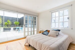 Quarto com cama, mesa de cabeceira, janela e vista para um pátio externo com plantas e cadeiras.