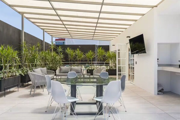 Um pátio externo moderno com mesa de vidro e cadeiras brancas, rodeado por plantas em vasos e uma TV fixada na parede.
