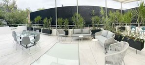 Um pátio externo moderno com mesa de vidro, cadeiras brancas, um sofá e plantas em vasos.