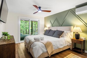 A bedroom with a bed, bedside lamps, a ceiling fan, a TV, and a window with a view of trees. - 7BR Fully Renovated Home w/ Hot Tub, Mini Golf. Sleeps 18! (McGaheysville)