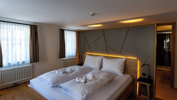 A hotel room with a large bed, a bedside table, a radiator, and a window with curtains. - Münsterhus Josefs Alpine Lodge - Zimba four-bed room (Schruns)