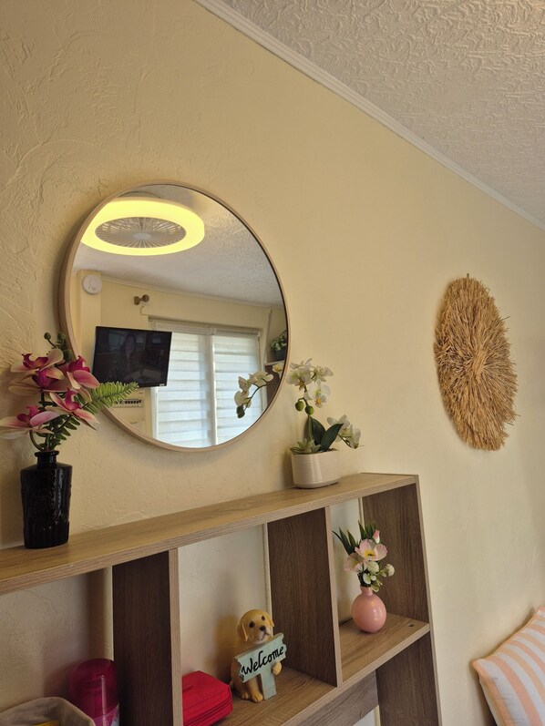 Una habitación con un espejo redondo, un televisor, un estante de madera con objetos decorativos y un elemento texturizado en la pared.