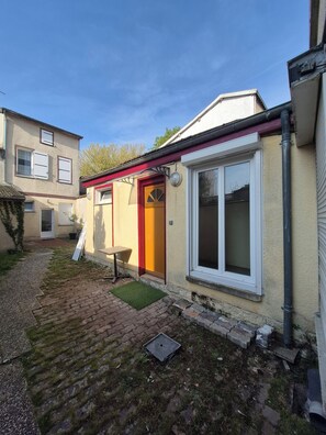 Un edificio residencial con puerta roja, ventana con marcos blancos y un pequeño espacio al aire libre con asientos.