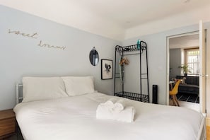 Une chambre avec un lit, une table de chevet, un miroir fixé au mur, un lit superposé en métal noir et une porte donnant sur une autre pièce.