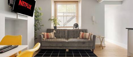 A living room with a grey sofa, a black rug, a yellow chair, a TV mounted on the wall, and a window with blinds.