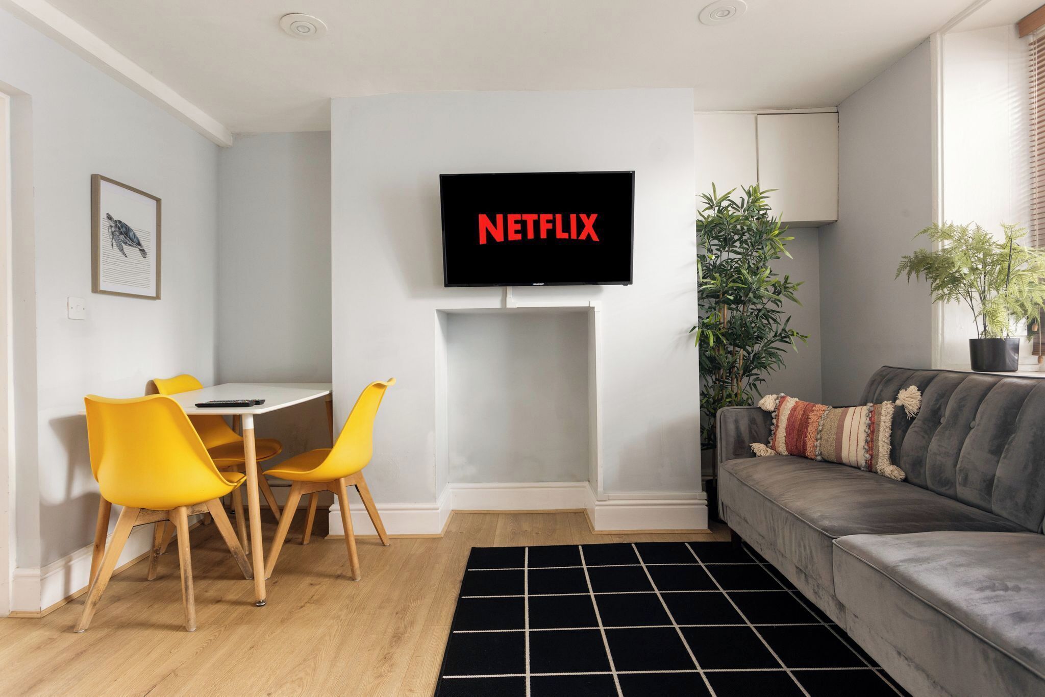 A living room with a TV mounted on the wall, a gray sofa, yellow chairs, a small table, and a black and white rug.