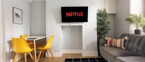 A living room with a TV mounted on the wall, a gray sofa, yellow chairs, a small table, and a black and white rug.