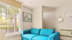 Una habitación con un sofá azul, un ventanal con vista a follaje, un cuadro enmarcado en la pared y un reloj fijado a la pared.