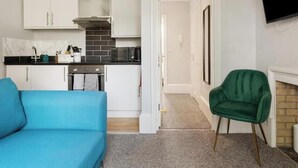 Un espacio habitable compacto con kitchenette, un sofá azul, un sillón verde y una chimenea.