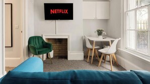 Una sala de estar con chimenea, un sillón verde, una mesa blanca y un televisor en la pared mostrando 'NETFLIX'.