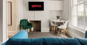 Ein Wohnzimmer mit Kamin, einem grünen Sessel, einem weißen Tisch und einem an der Wand befestigten Fernseher, der 'NETFLIX' zeigt.