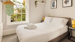 Una cama bien hecha con sábanas blancas, una mesita de noche con una lámpara amarilla y una ventana con vistas al verdor exterior.