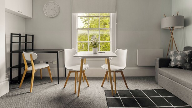 Ein modernes Zimmer mit Esstisch, zwei Stühlen, einer Couch, einer Lampe und einem Fenster mit Blick auf Grünflächen.