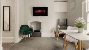 Ein modernes Wohnzimmer mit einem Fernseher, der 'NETFLIX' zeigt, einem grünen Sessel, einem weißen Esstisch und einer Topfpflanze.