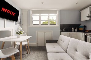 Eine moderne Küche mit Einbauofen, ein Essbereich mit weißem Tisch und Stühlen, ein graues Sofa und ein Fenster mit Blick auf Grünflächen.