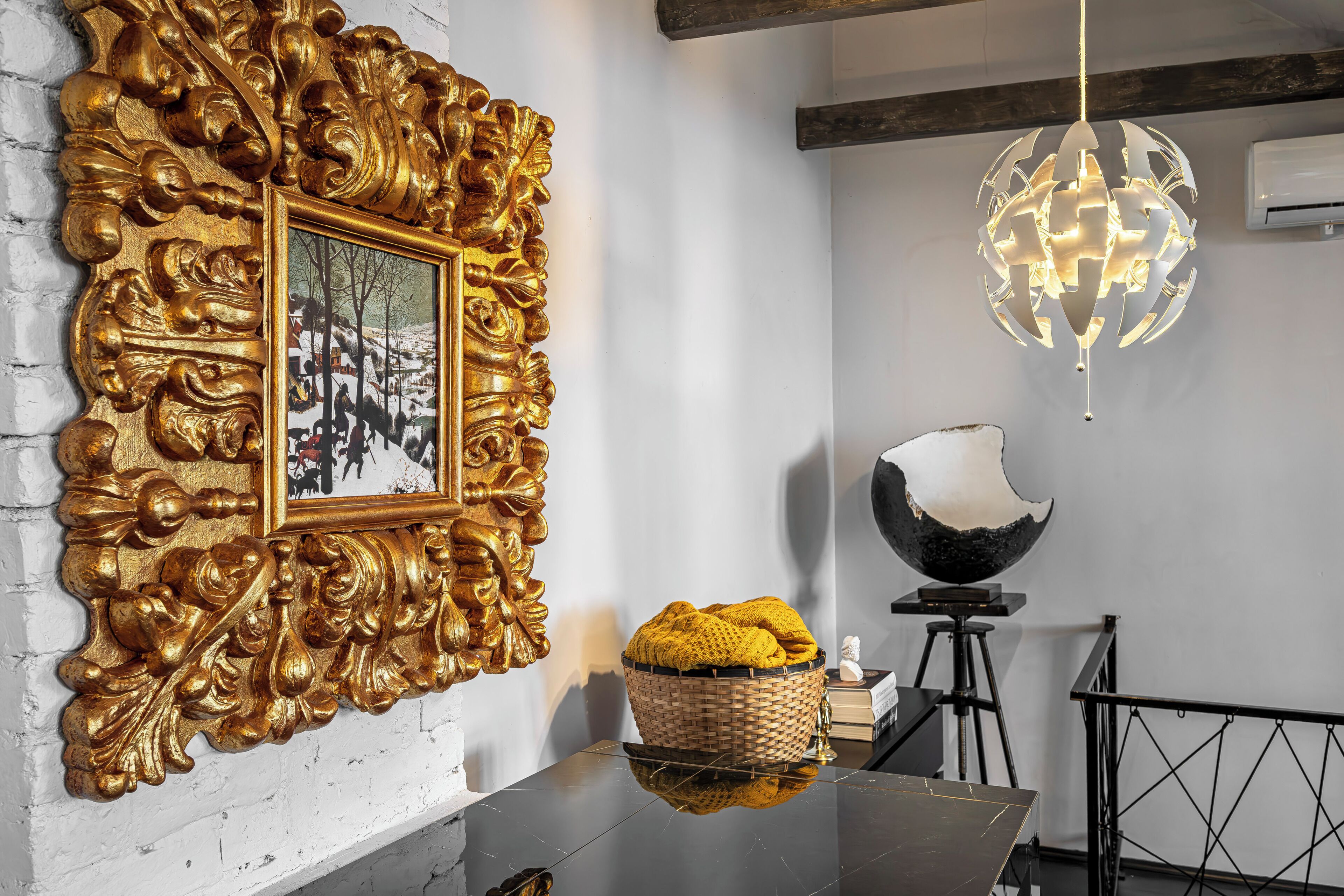 A room with a gold ornate mirror, a framed picture, a reflective black table, a woven basket, a modern spherical light fixture, and a black tripod stand.