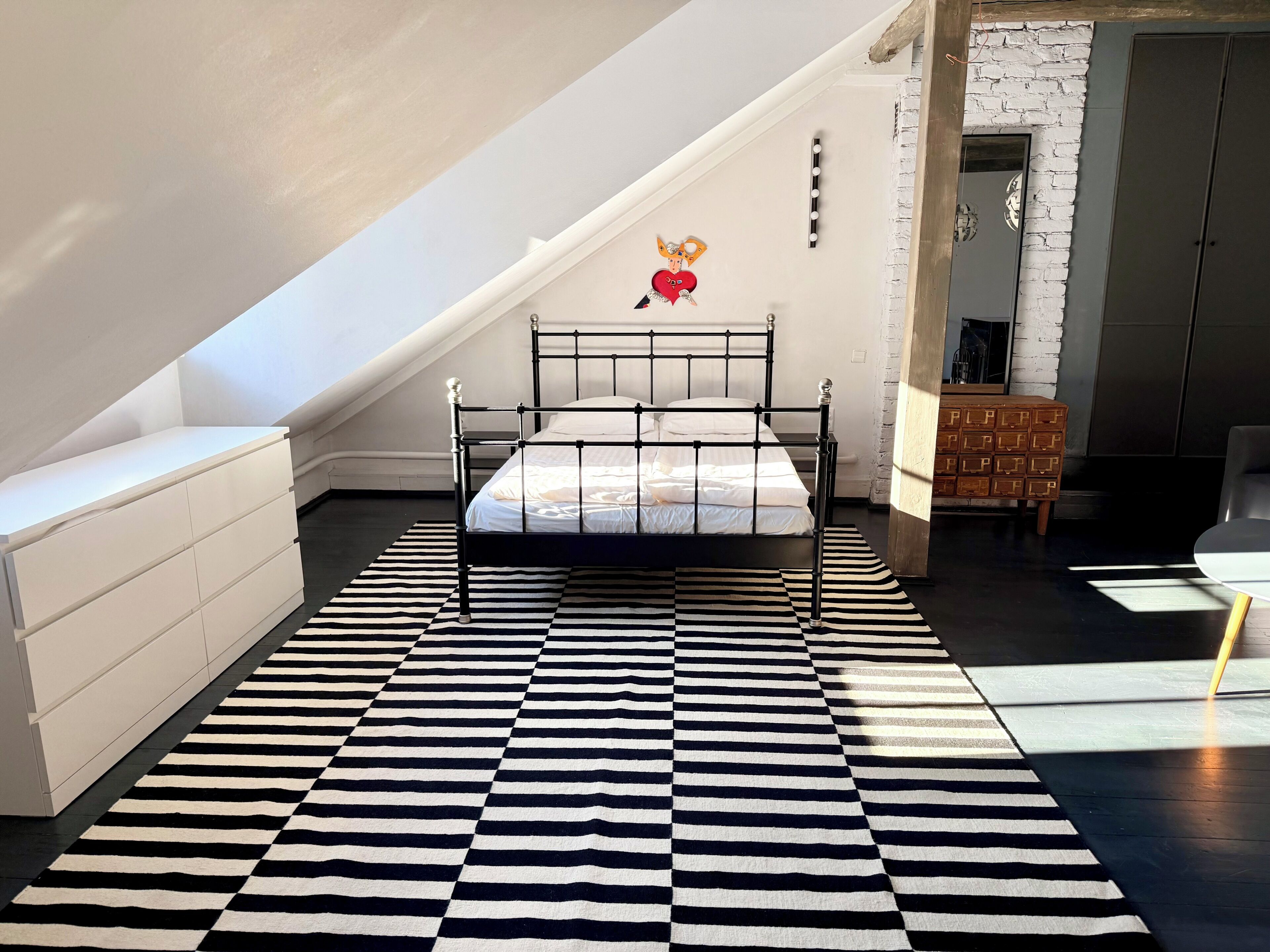 Ein Schlafzimmer mit einem schwarz-weißen StreifenTeppich, einem Bett, einer weißen Kommode und einer Ziegelwand.