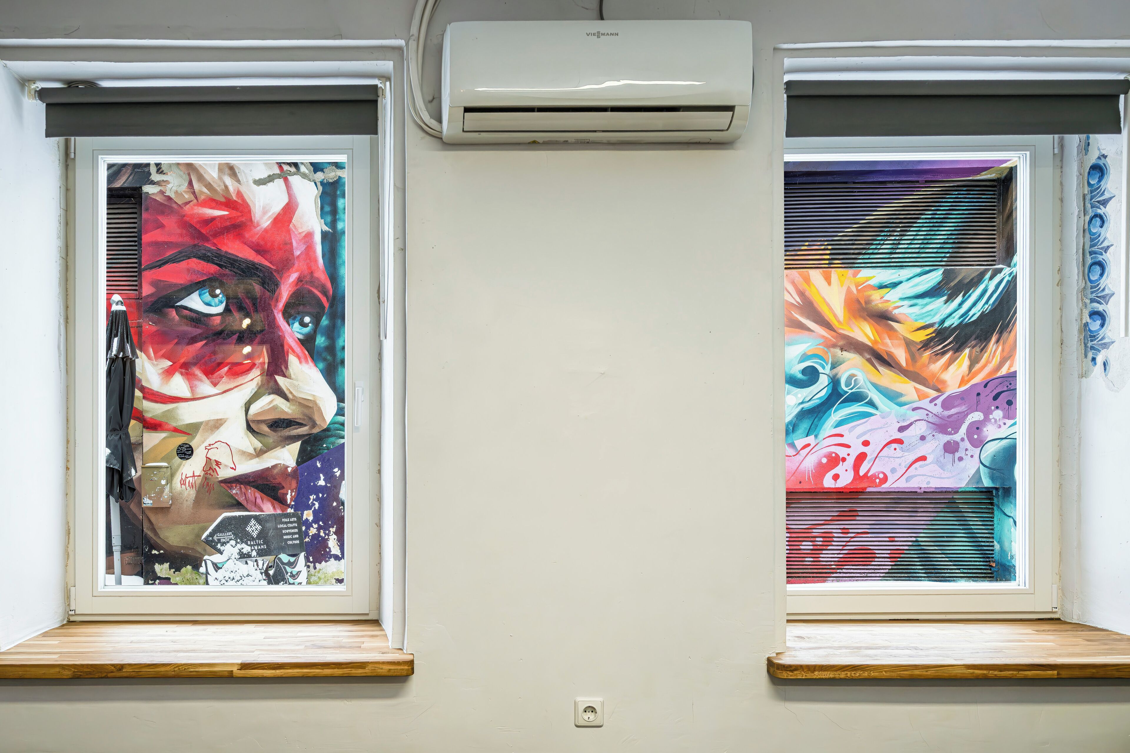 Dos ventanas con llamativos grafitis, una unidad de aire acondicionado blanca en la parte superior y un alféizar de madera.