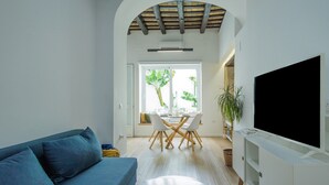 Een moderne woonkamer met een bank, een eettafel met stoelen, een flatscreen televisie en een groot raam met planten.