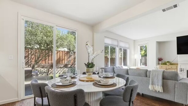 A dining area with a round table, grey chairs, and a view of the backyard through large sliding glass doors.