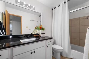 A bathroom with a black countertop, white cabinets, a mirror with lights, a toilet, and a shower area.