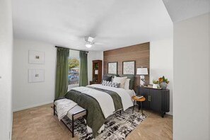 Une chambre à coucher comprenant un lit, une table de chevet, une commode, un ventilateur de plafond et une fenêtre avec des rideaux.