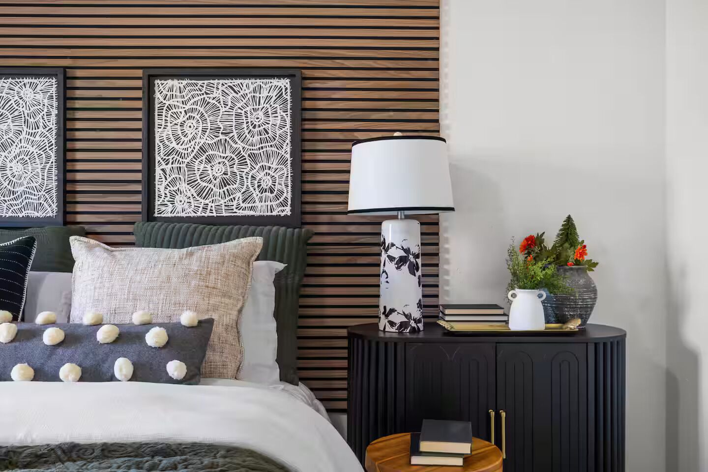 A neatly arranged bedroom with a bed, pillows, a bedside table with a lamp, a vase with flowers, and a wall with patterned panels.