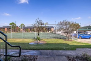 Un court de tennis entouré d’une clôture, avec un petit arbre et une zone de stationnement où sont garées des voitures.