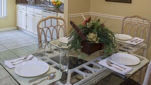 Un comedor con una mesa de vidrio dispuesta para cuatro personas, rodeada de sillas, un centro decorativo y una cocina al fondo.