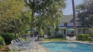 Una piscina con sillas de descanso y un pequeño jacuzzi, rodeado de árboles y un edificio.