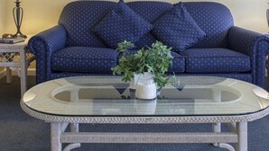 Una habitación con un sofá azul, una mesa de centro de vidrio, un cuadro en la pared y dos lámparas sobre mesitas auxiliares.