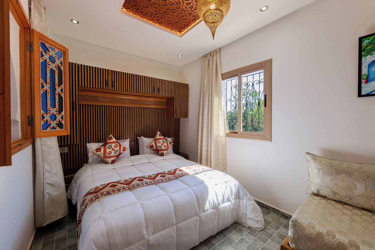 A bedroom with a bed, a window with a view, a decorative ceiling, and a wall-mounted shelf.