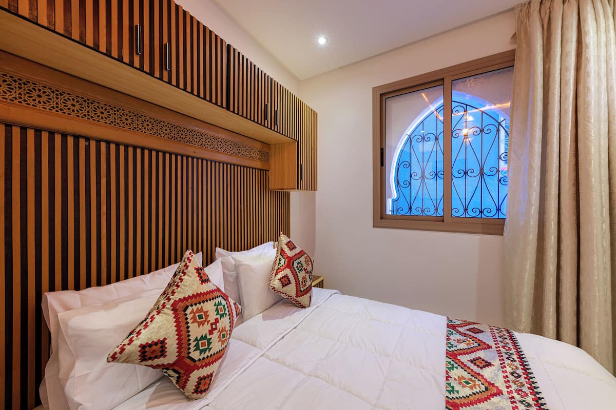 A hotel room with a bed, pillows, a window with a metal gate, and a wooden headboard.