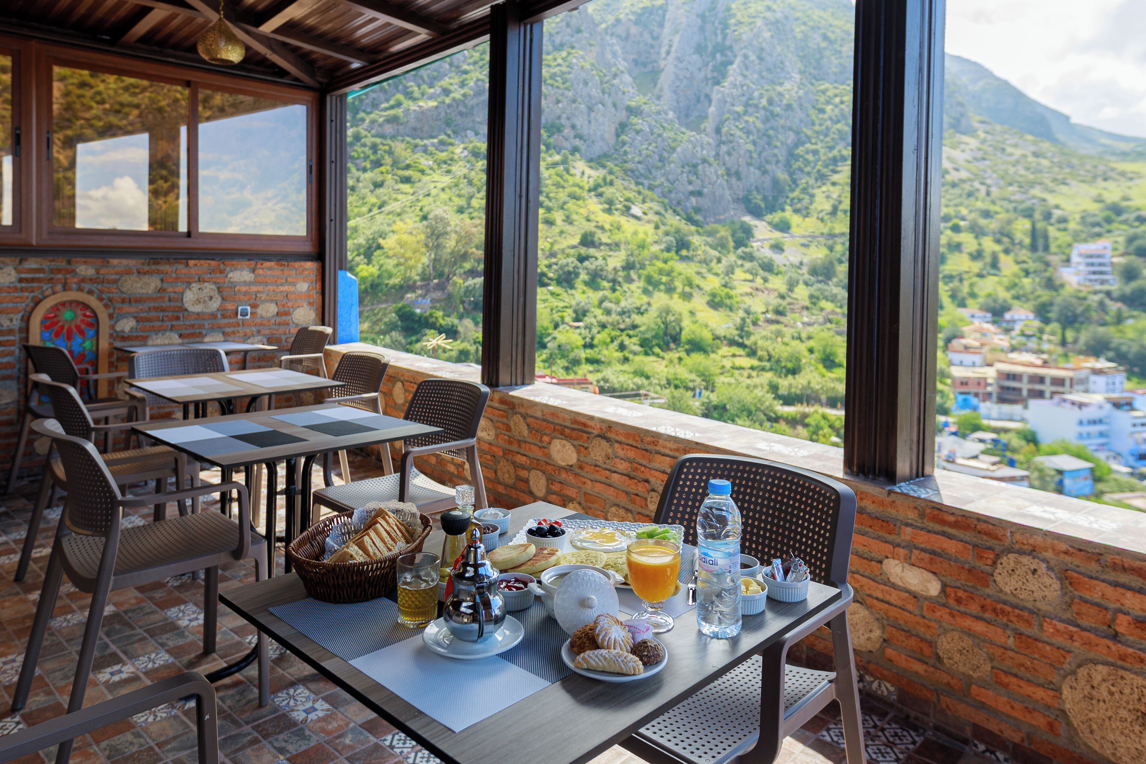 Un espace extérieur aménagé pour les repas, avec des tables et des chaises, un panier de pain, une théière et une vue sur un paysage montagneux.