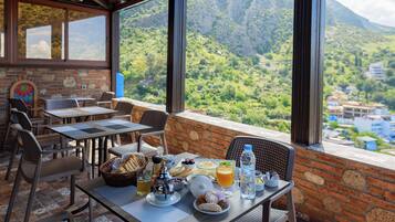 Un espace extérieur aménagé pour les repas, avec des tables et des chaises, un panier de pain, une théière et une vue sur un paysage montagneux.